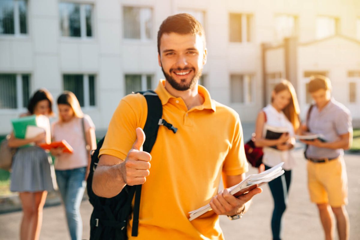 Jovem universitário posa sorridente fazendo sinal de positivo com olegar para cima no campus da universidade com outros alunos ao fundo