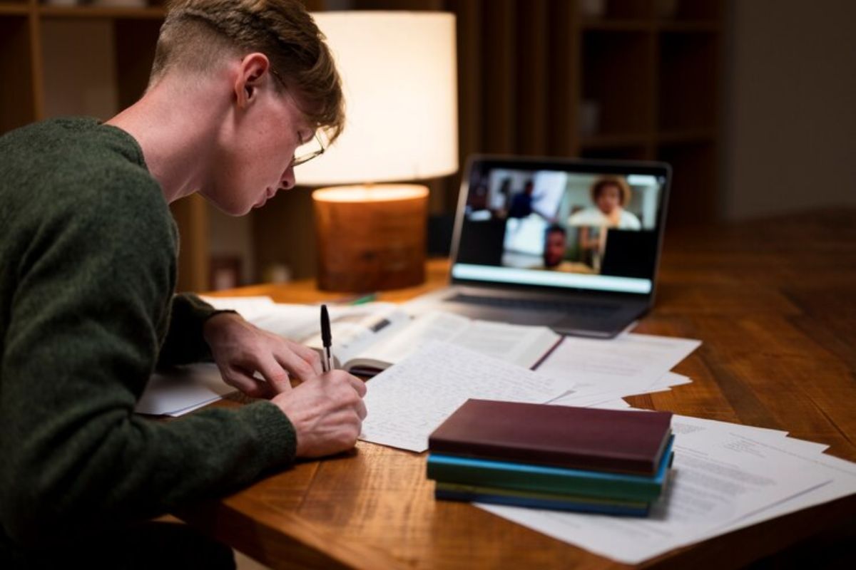 Um jovem é visto de costas em seu quarto. Ele está estudando, enquanto vê uma videoaula, faz anotações em seu caderno 