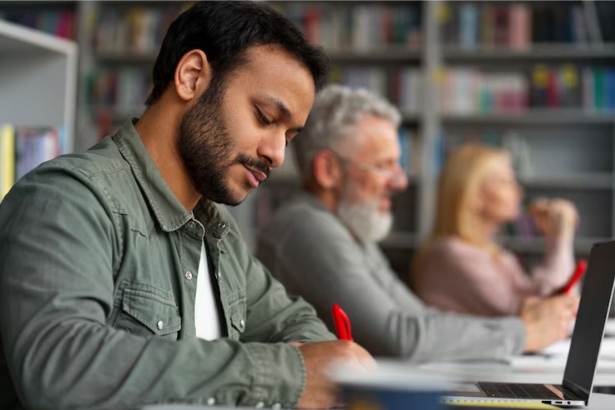 Um jovem em frente ao seu notebook se inscreve nos cursos EAD SISU 2025. Ele é visto de lado, dentro de uma sala de aula