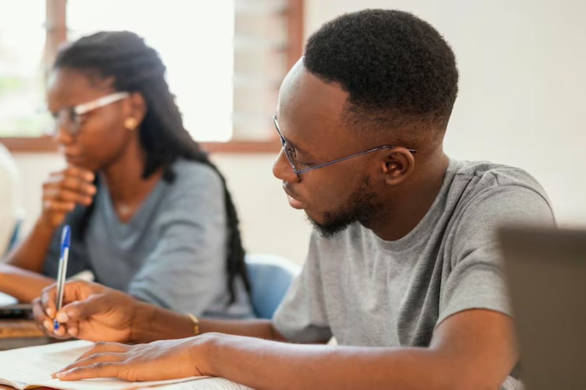 Dois estudantes em uma sala de aula. O foco é em um jovem que escreve em seu caderno