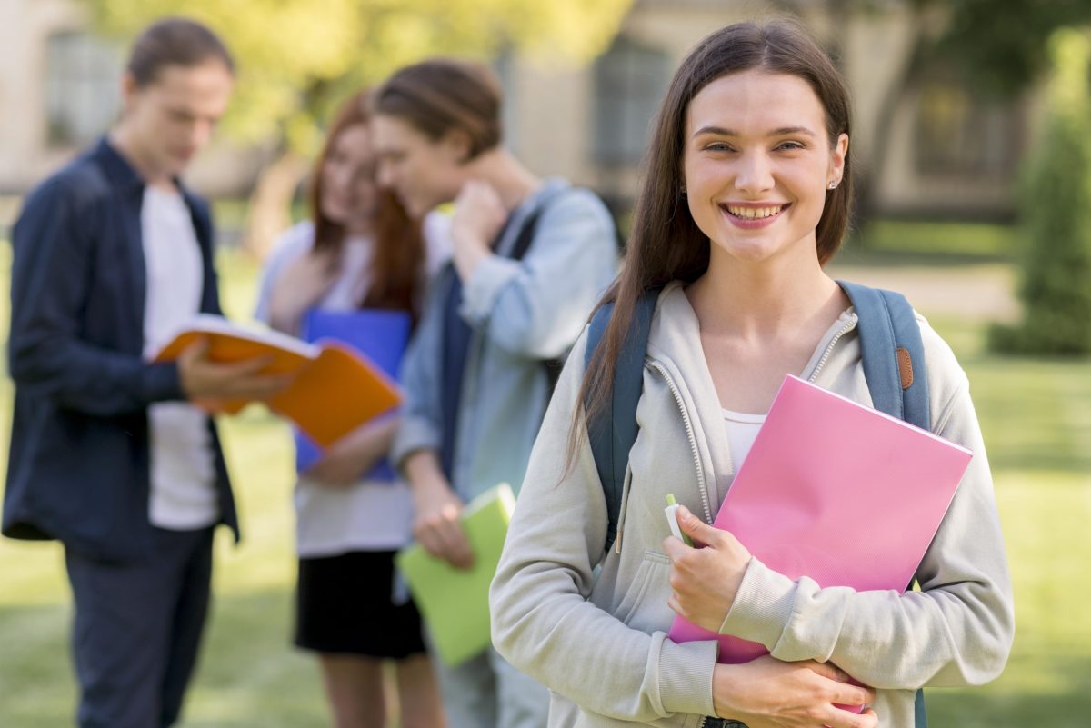 jovem universitária sorridente posa carregando materiais nas mãos com colega ao fundo no campus