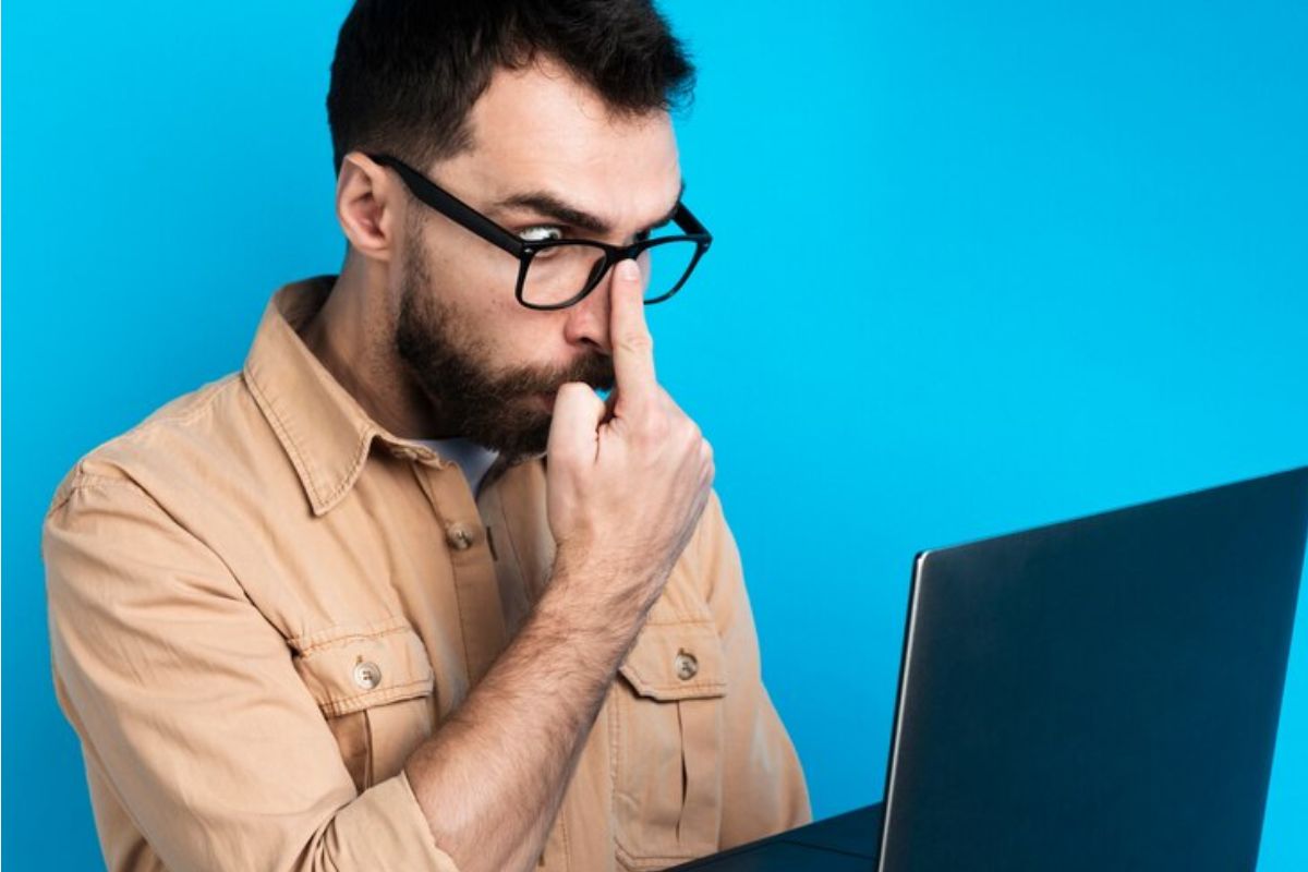 Um homem ajeita o seu óculos, ele segura o notebook enquanto está de pé, pesquisando os cursos com notas de corte mais baixas de toda história do SISU