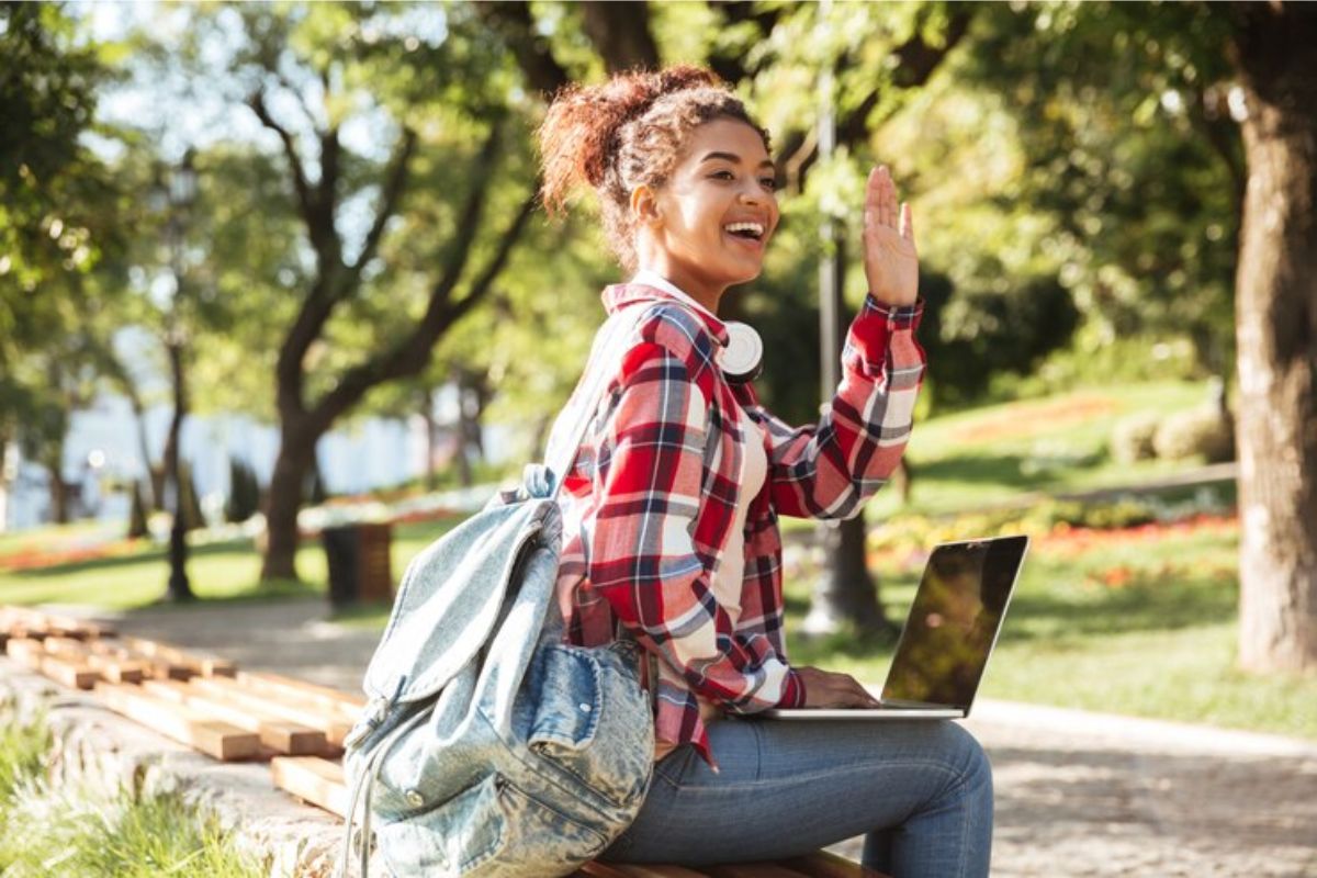 Uma estudante no jardim da universidade sentada em um banco com o notebook no colo. Ela acena para alguém que vai embora