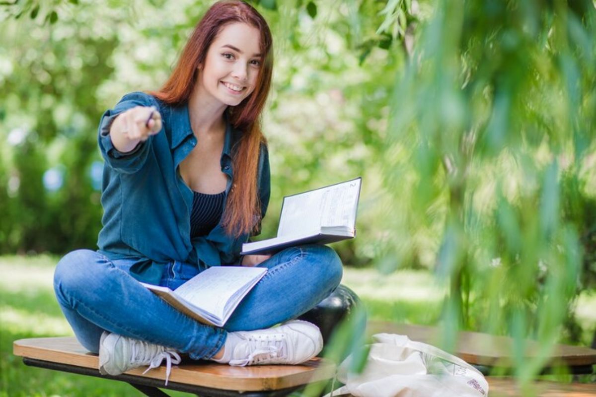 Uma estudante sentada no jardim da faculdade. Ela segura um livro e aponta para a câmera com um lápis sorrindo