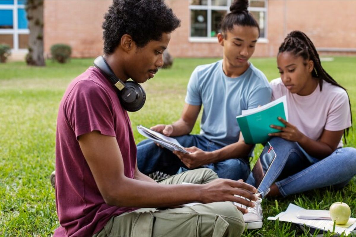 Três estudantes sentados na grama estudam juntos. Um usa o seu notebook no colo enquanto outros dois colegas conversam com um livro aberto na mão