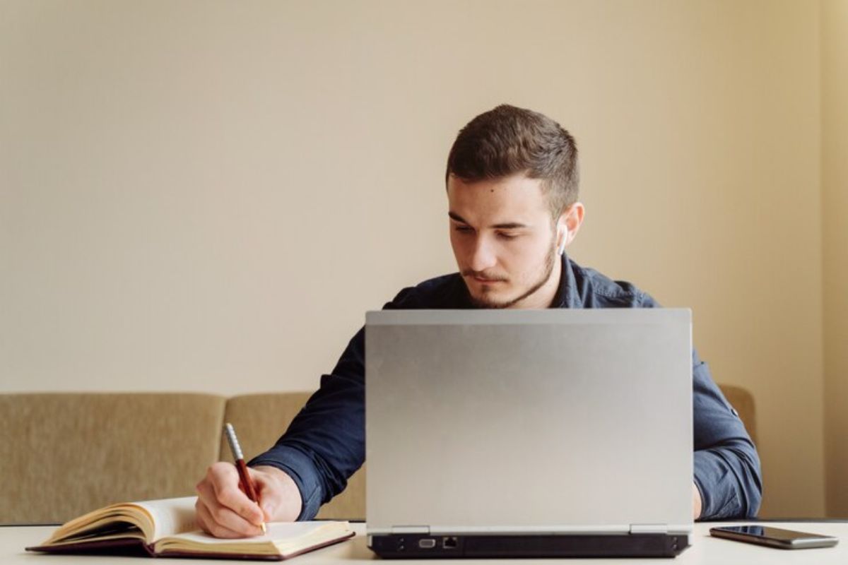 Um jovem que quer passar no SISU faz anotações em seu caderno enquanto consulta as menores notas em seu notebook