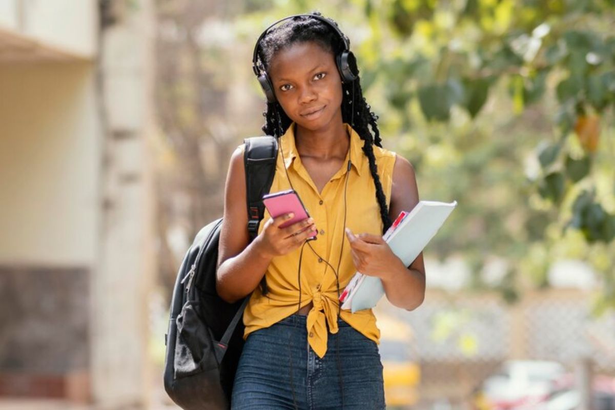 Uma estudante segura parte de seu material em um braço e olha o celular na outra mão