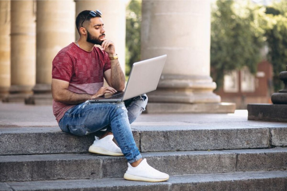 Um estudante com o notebook no colo em uma escadaria. Ele pensa bem antes de fazer sua escolha pois Quer passar no SISU