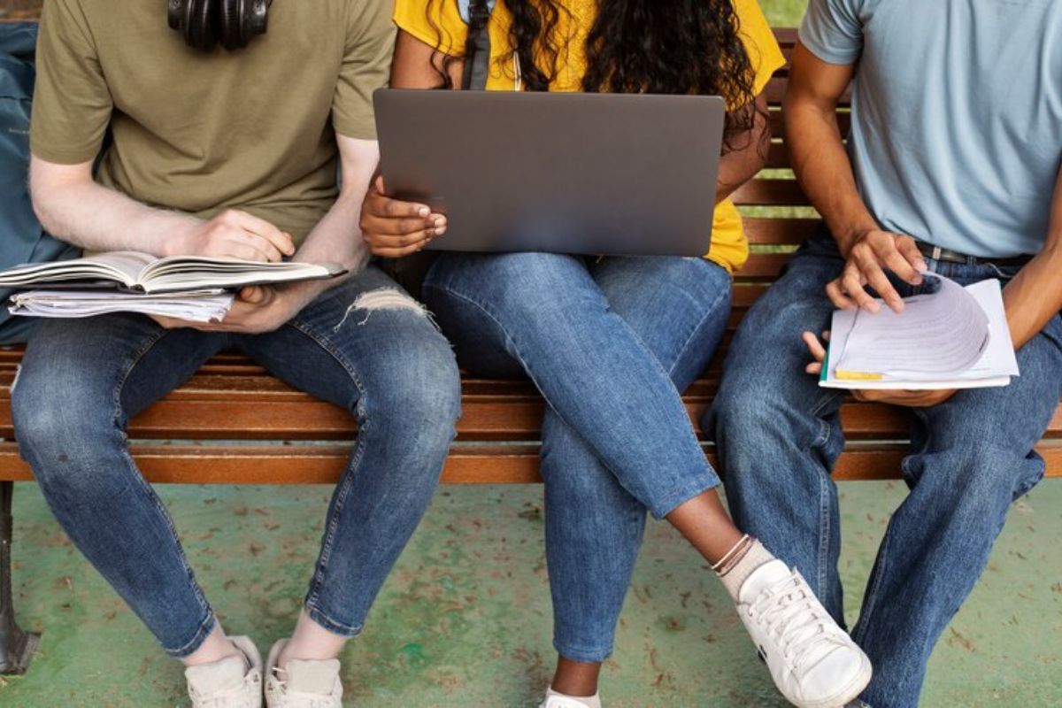 Três estudantes sentados em um banco. Uma aluna no meio segura um notebook onde os estudantes conferem o edital do SiSU